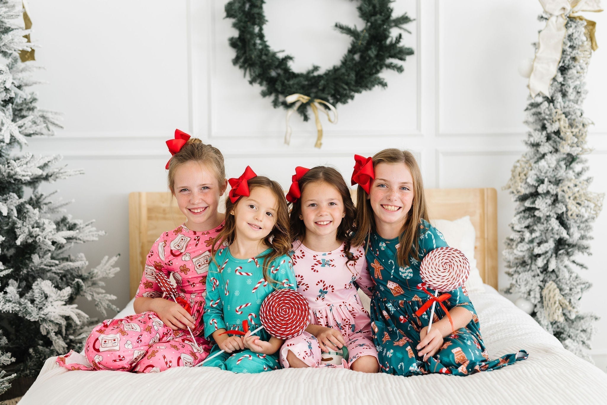 Four smiling girls in festive holiday dresses and red hair bows holding lollipops on a bed with Christmas decor.