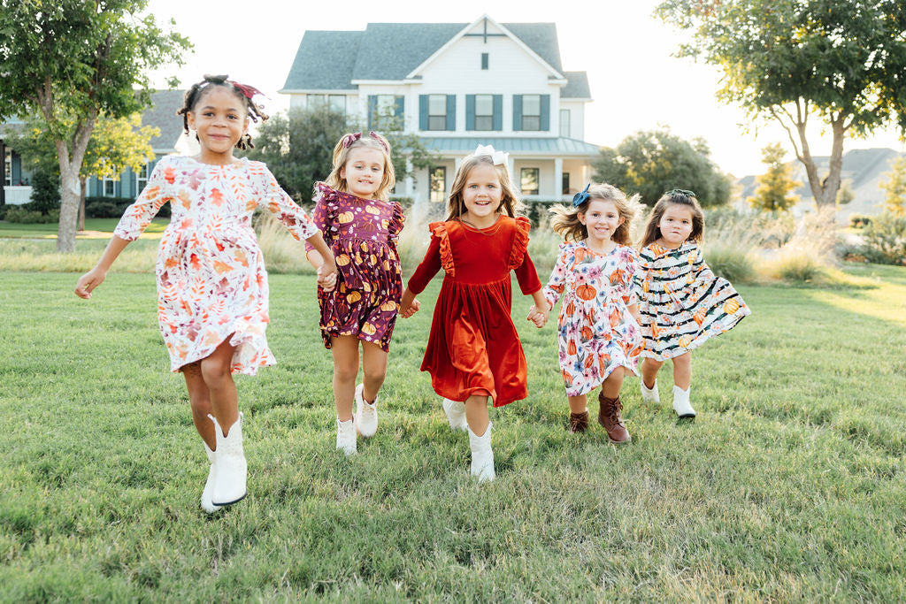 Five young girls in festive fall dresses and cowboy boots running together on a grassy lawn for Thanksgiving.