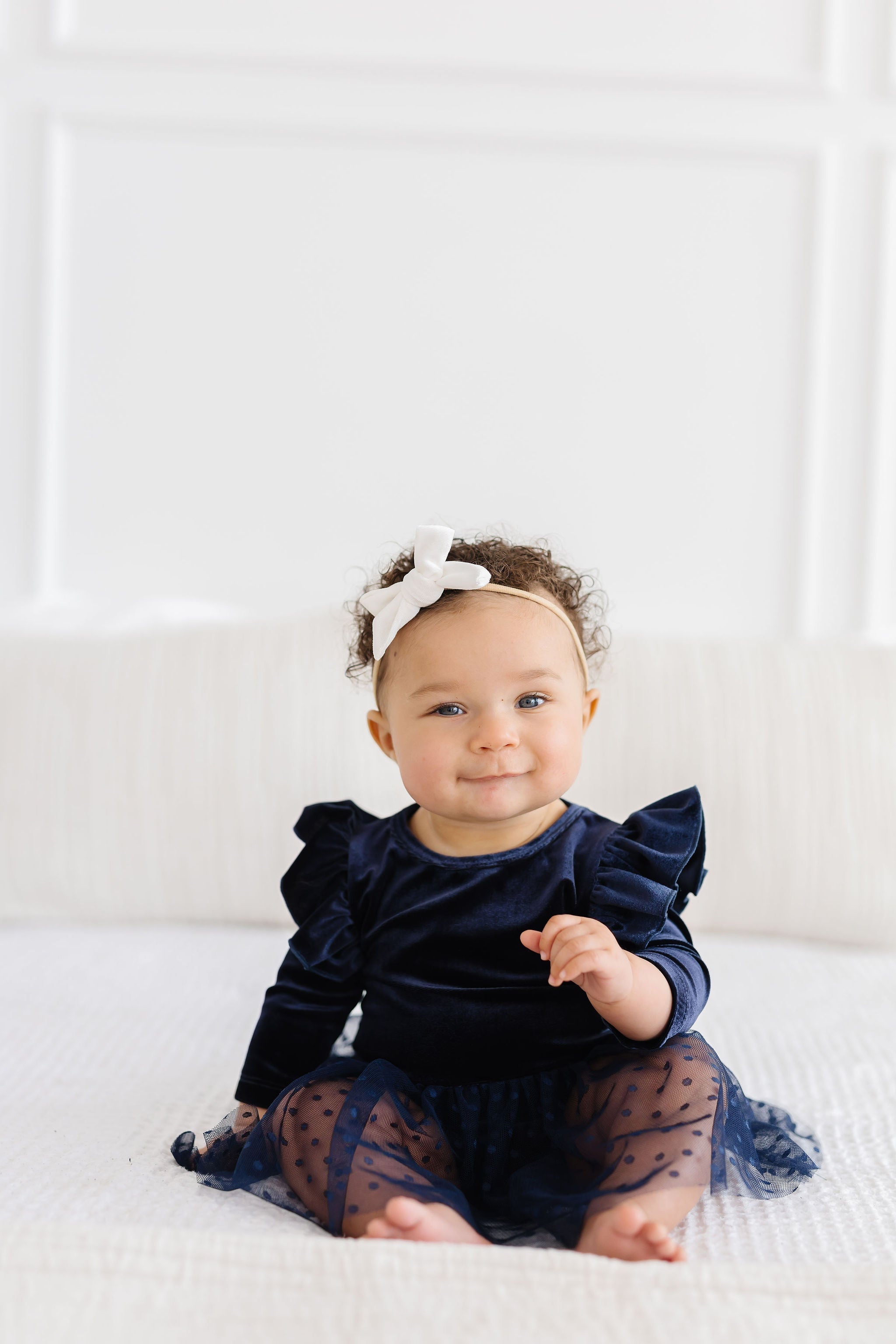 Baby girl wearing a soft, navy blue velvet dress with a tulle skirt and a white bow headband on a white bed.