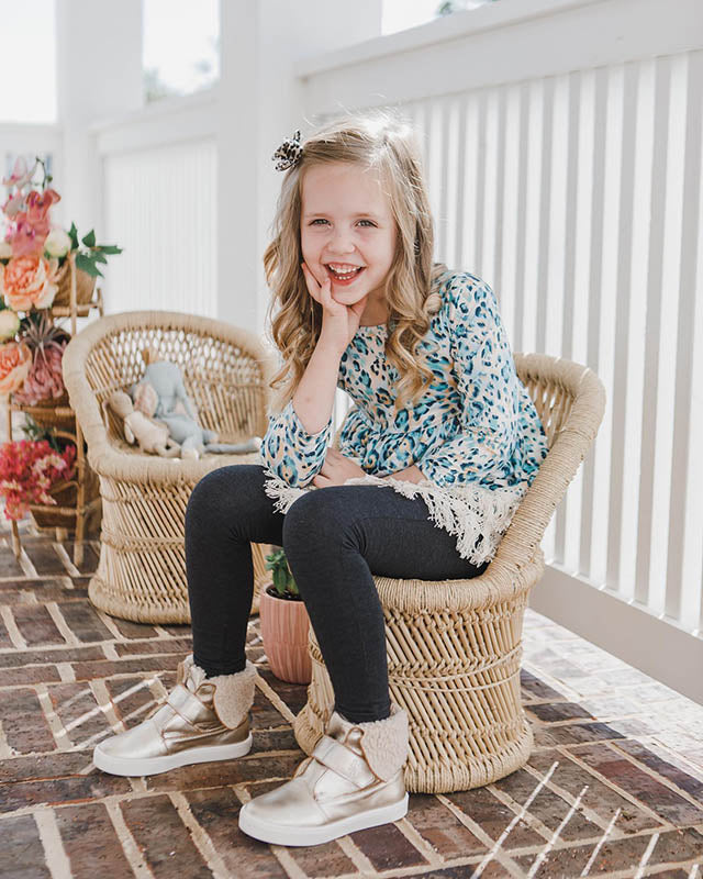 Young girl in a boho leopard print peplum top, dark leggings, and metallic boots sitting on a wicker chair.