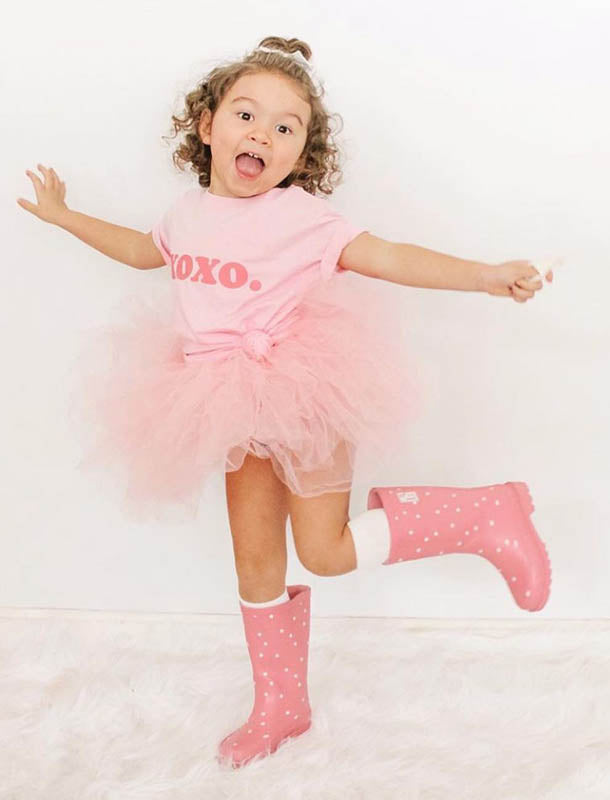 Happy toddler wearing a pink XOXO t-shirt, fluffy tulle skirt, and polka-dot rain boots jumping joyfully.