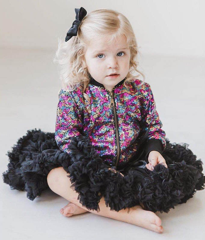 Toddler girl wearing a vibrant multicolored sequin bomber jacket with a voluminous black ruffled tulle skirt.