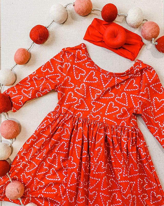 Red long-sleeve girls' twirl dress with candy cane heart print and matching red headband, accented by felt ball garland.