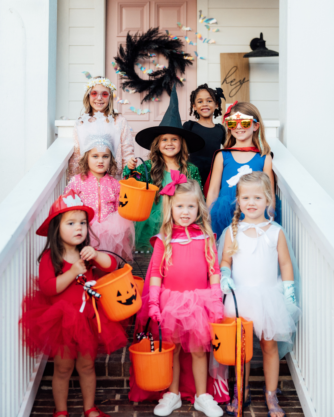 Group of young girls in colorful Halloween costumes holding orange jack-o'-lantern candy buckets on a porch.