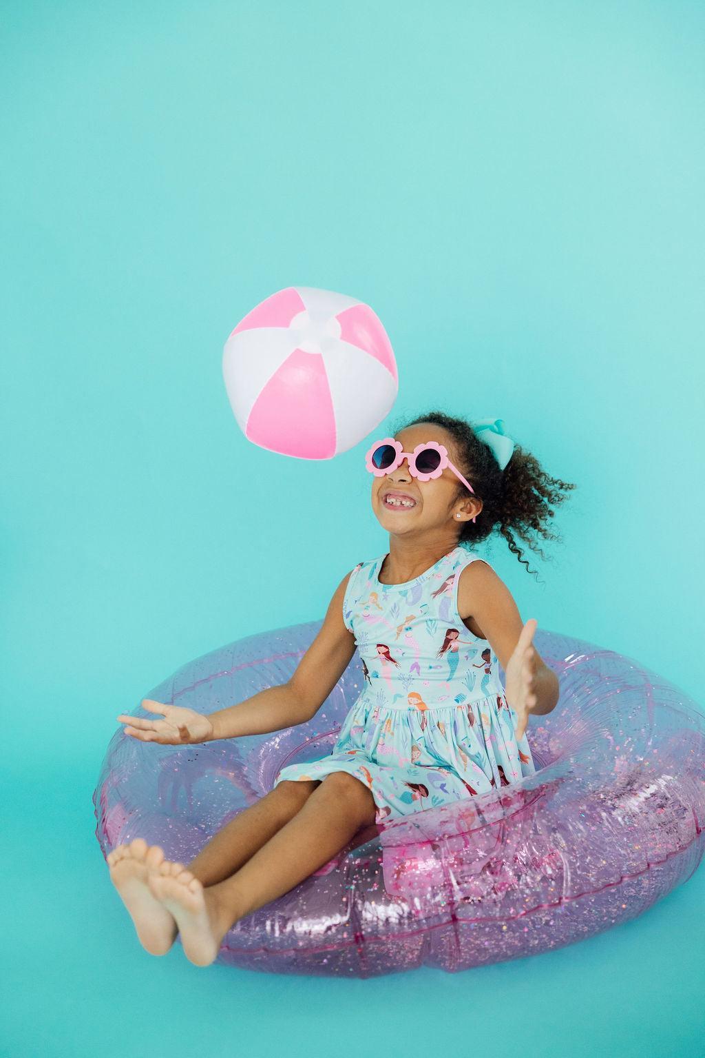 Young girl in a mermaid dress and flower sunglasses playing with a pink and white beach ball in a glittery pool float.