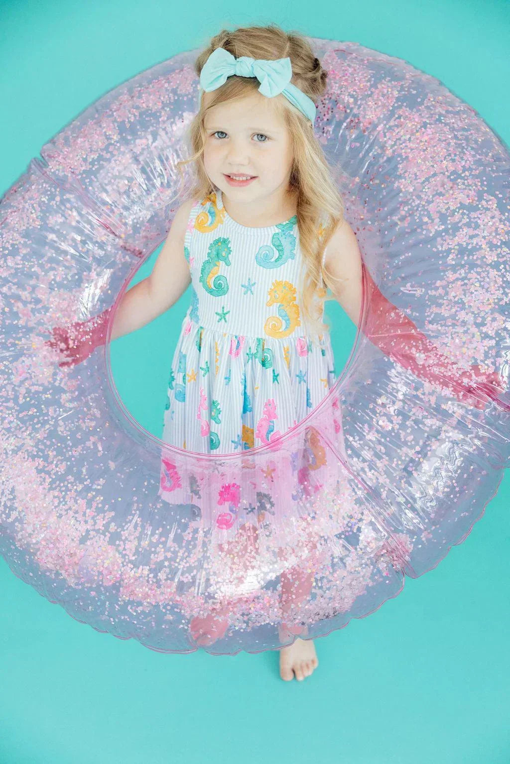 Young girl in a seahorse-print dress holding a large, clear inflatable pool ring filled with pink glitter.