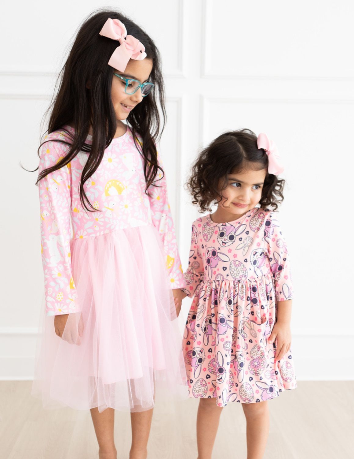 Two young girls holding hands, wearing pink Easter-themed dresses with bunny and floral patterns and matching hair bows.