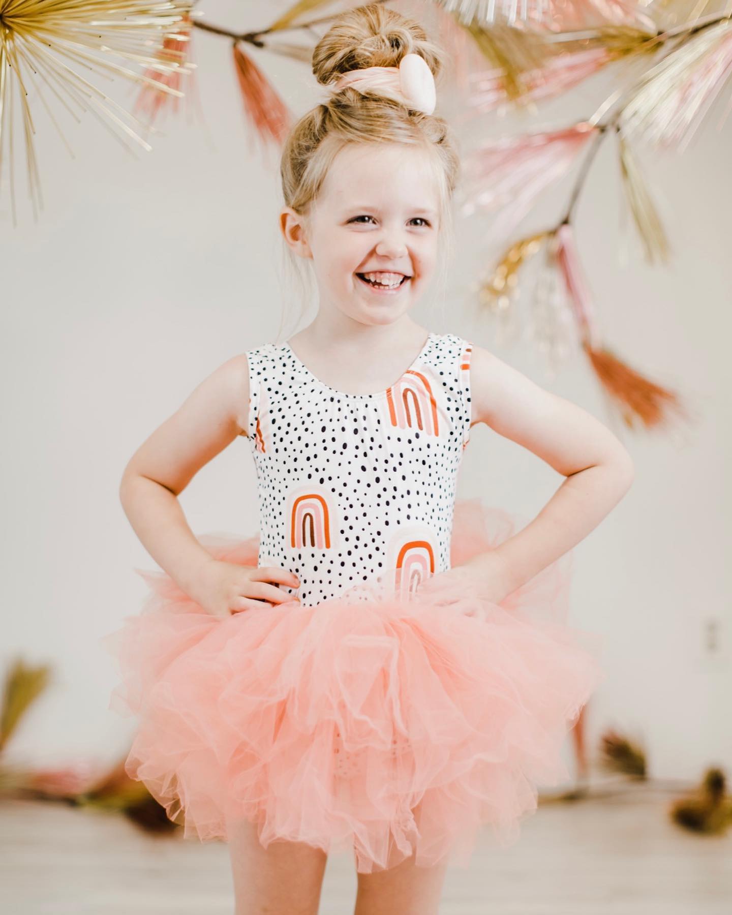 Little girl smiling in a white leotard with rainbow print and a fluffy peach tulle tutu skirt.