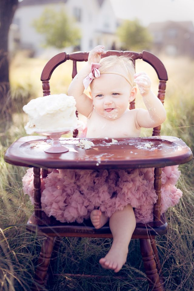 Baby girl in a pink tutu sitting in a wooden high chair, covered in frosting during a first birthday cake smash.