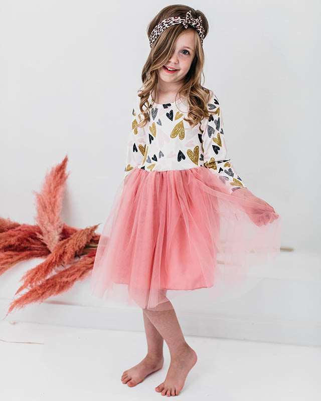 Little girl wearing a long-sleeve dress with gold and black hearts and a pink tulle skirt, styled with a leopard headband.