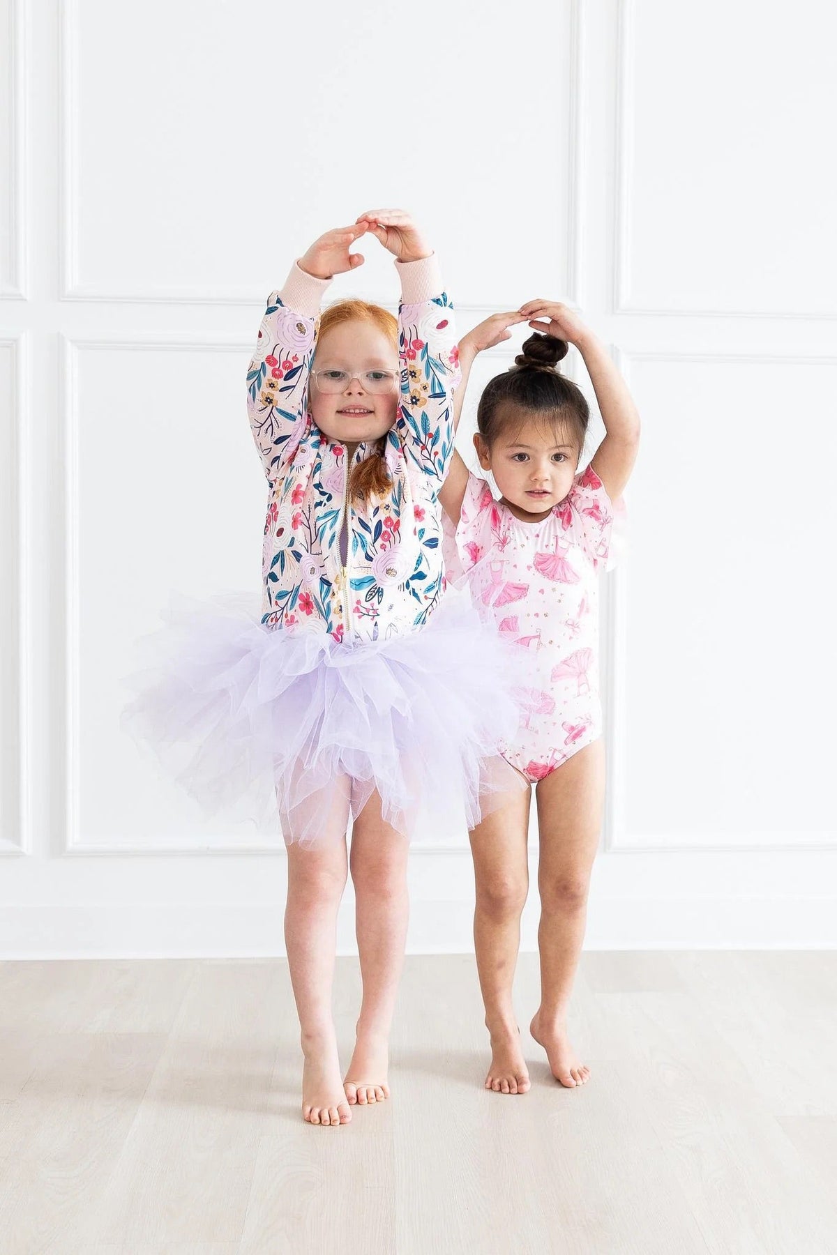 Two young girls posing in floral dancewear, featuring a lavender tulle tutu and a pink ballet-themed leotard.