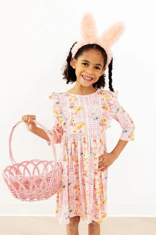 Smiling girl in a pink gingham chick-print dress wearing fluffy bunny ears and holding a pink wicker Easter basket.