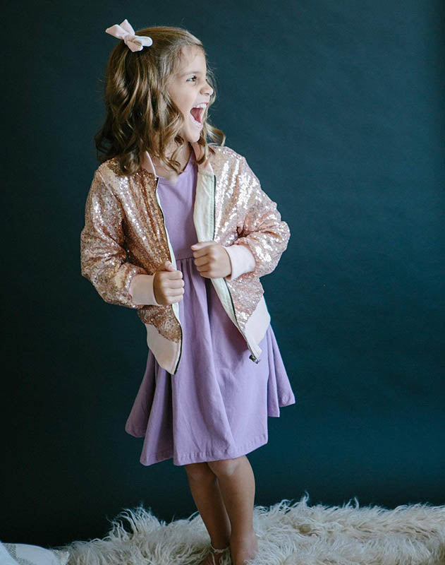 Happy young girl wearing a sparkling rose gold sequin bomber jacket over a simple purple dress.