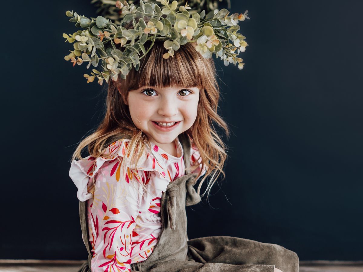 Young girl with bangs wearing a floral top and olive overalls, smiling while wearing a green eucalyptus crown.