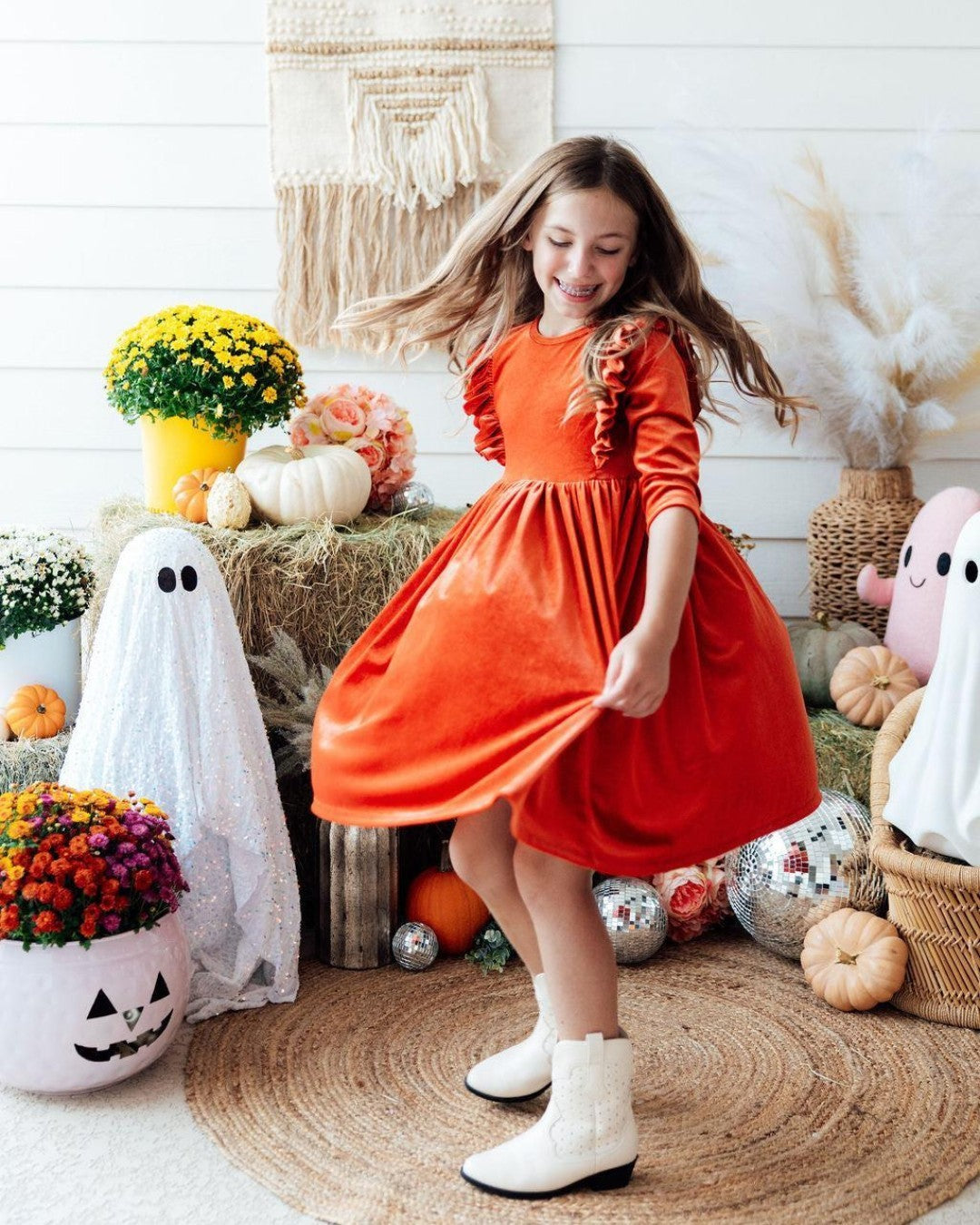 Girl twirling in a burnt orange velvet fall dress with ruffled sleeves and white ankle boots in a festive autumn setting.