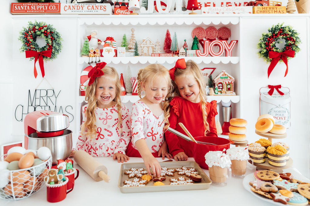 Three young girls in festive red outfits baking snowflake cookies in a bright, Christmas-decorated kitchen.