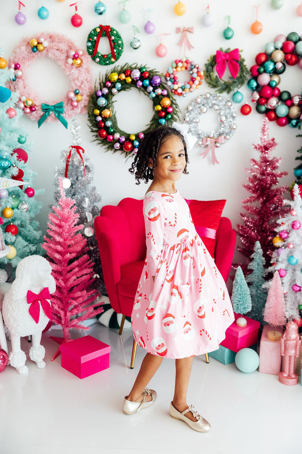 Young girl in a pink Santa-print dress standing in a festive room decorated with colorful Christmas trees and wreaths.