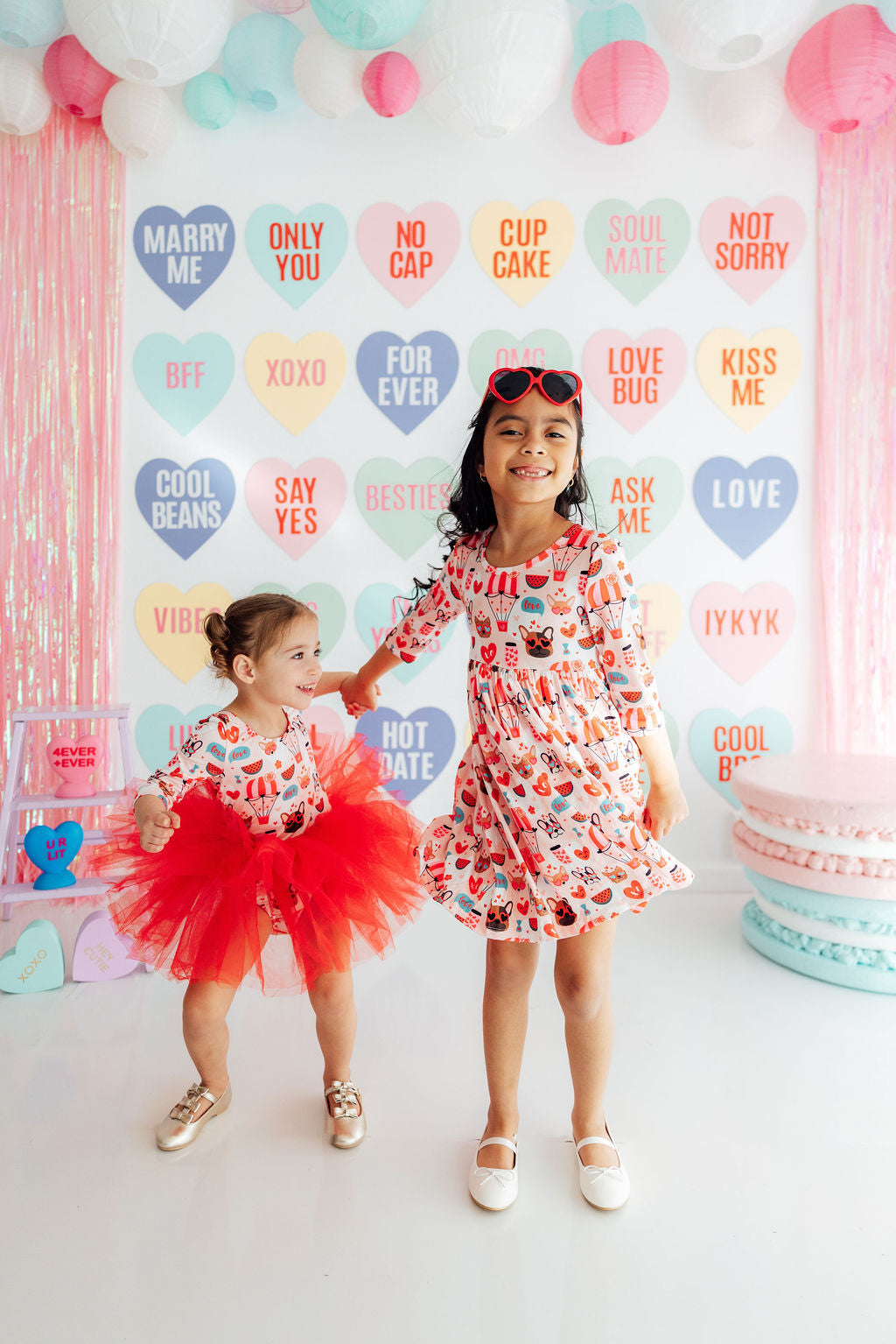 Young girl wearing a fluffy red tulle tutu and matching heart-print outfit standing next to her sister.