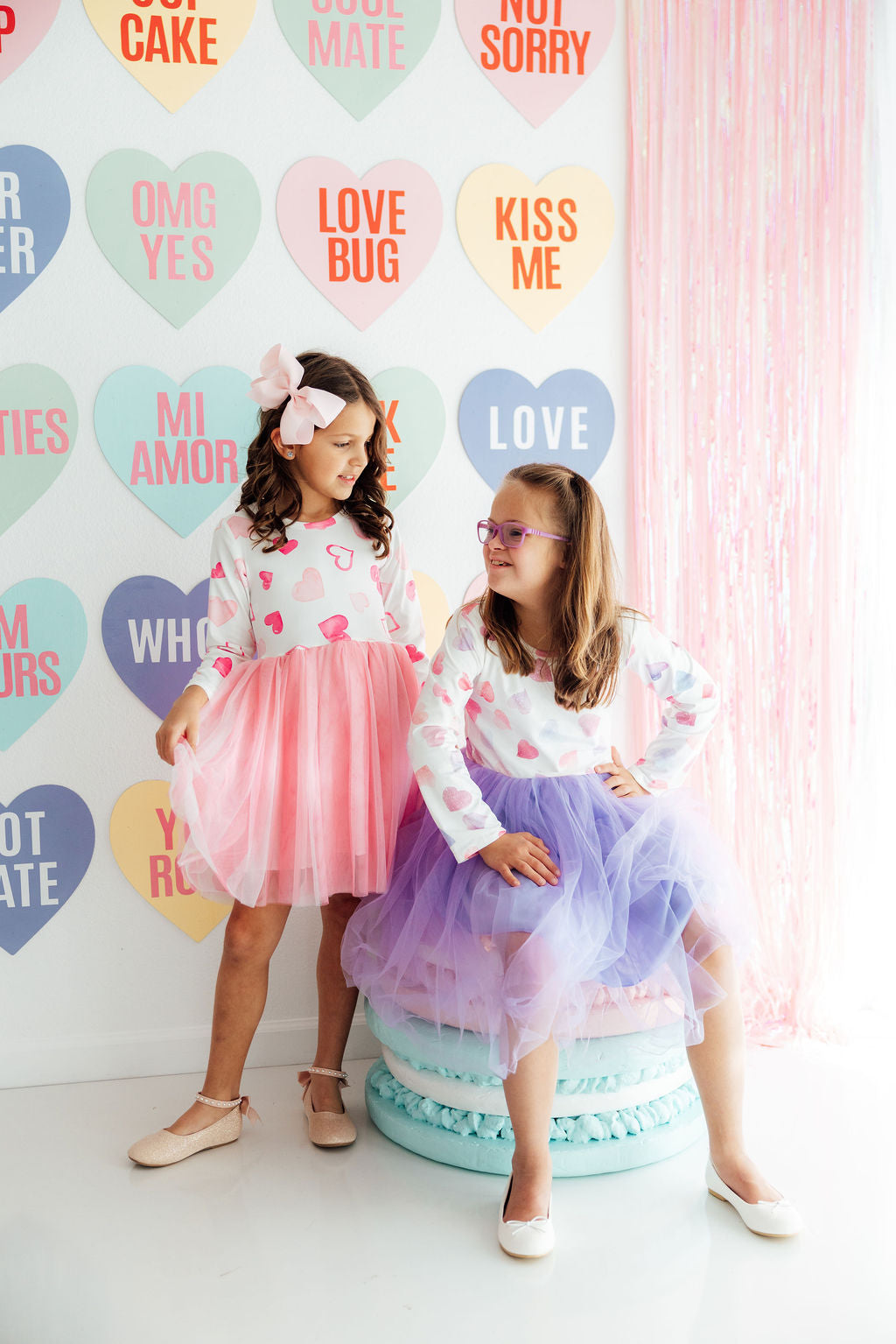 Two girls wearing heart-print long-sleeve dresses with pink and purple tulle skirts in front of a heart-themed wall.