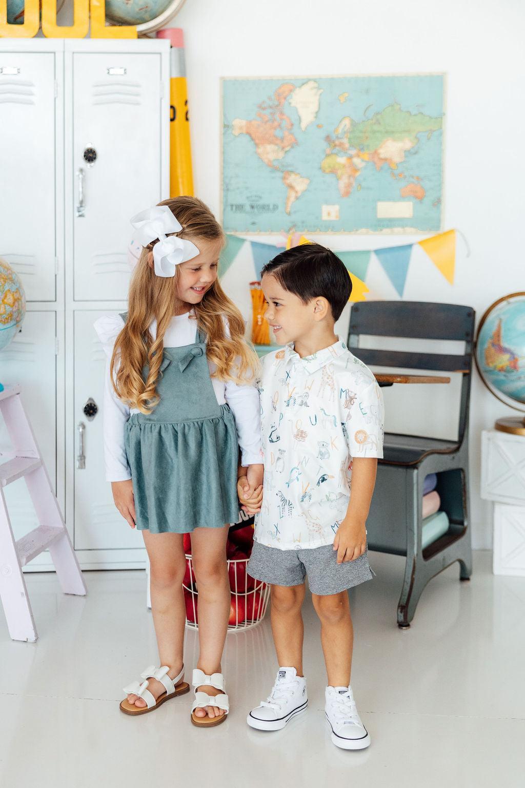 A young girl and boy holding hands in a classroom setting with a world map, lockers, and school supplies.
