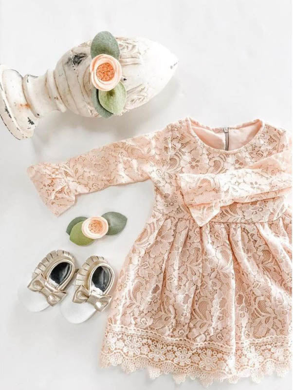 Blush pink lace baby dress paired with metallic gold moccasins and floral hair accessories on a white background.