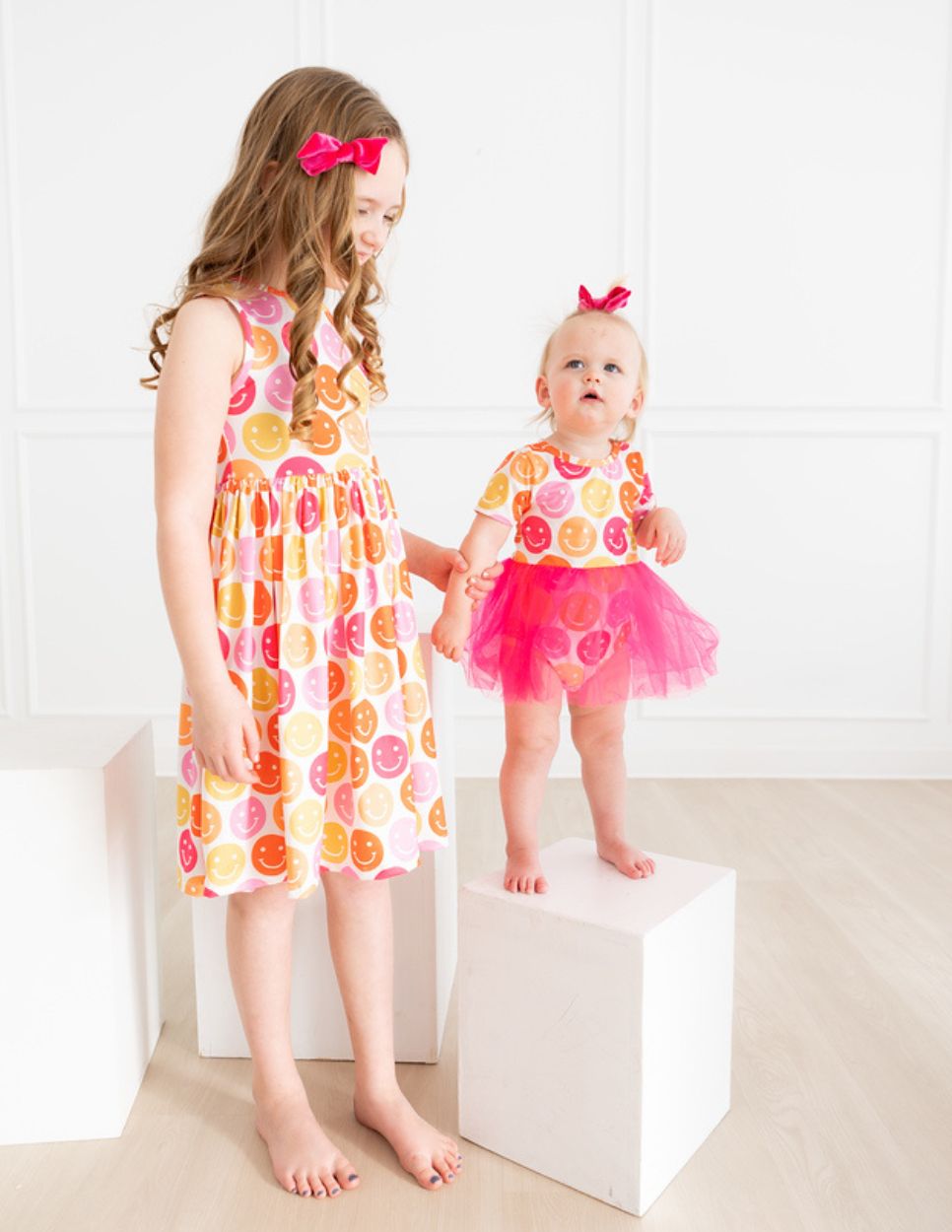 Two sisters wearing matching pink and orange smiley face outfits with coordinating velvet hair bows.