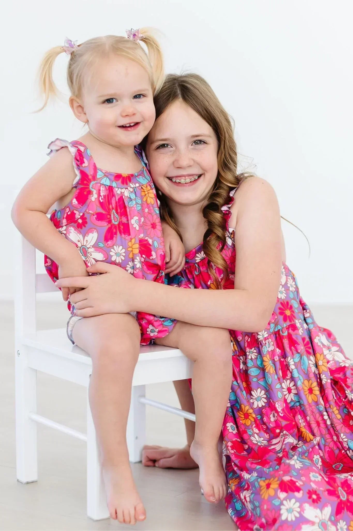 Two sisters smiling while wearing matching pink floral dresses with ruffled straps.