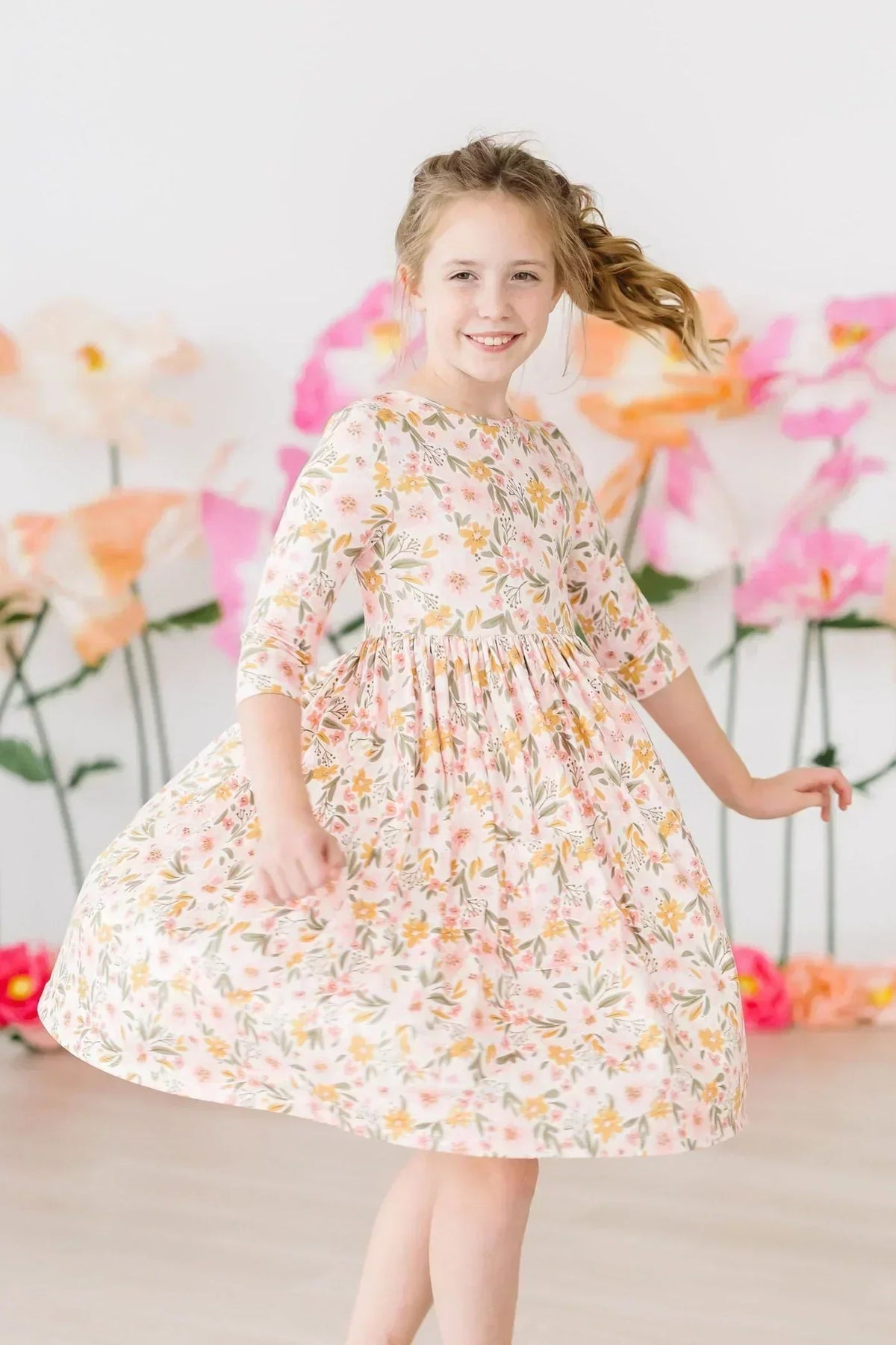 Young girl twirling in a soft pink floral Mila & Rose dress with three-quarter sleeves against a backdrop of paper flowers.