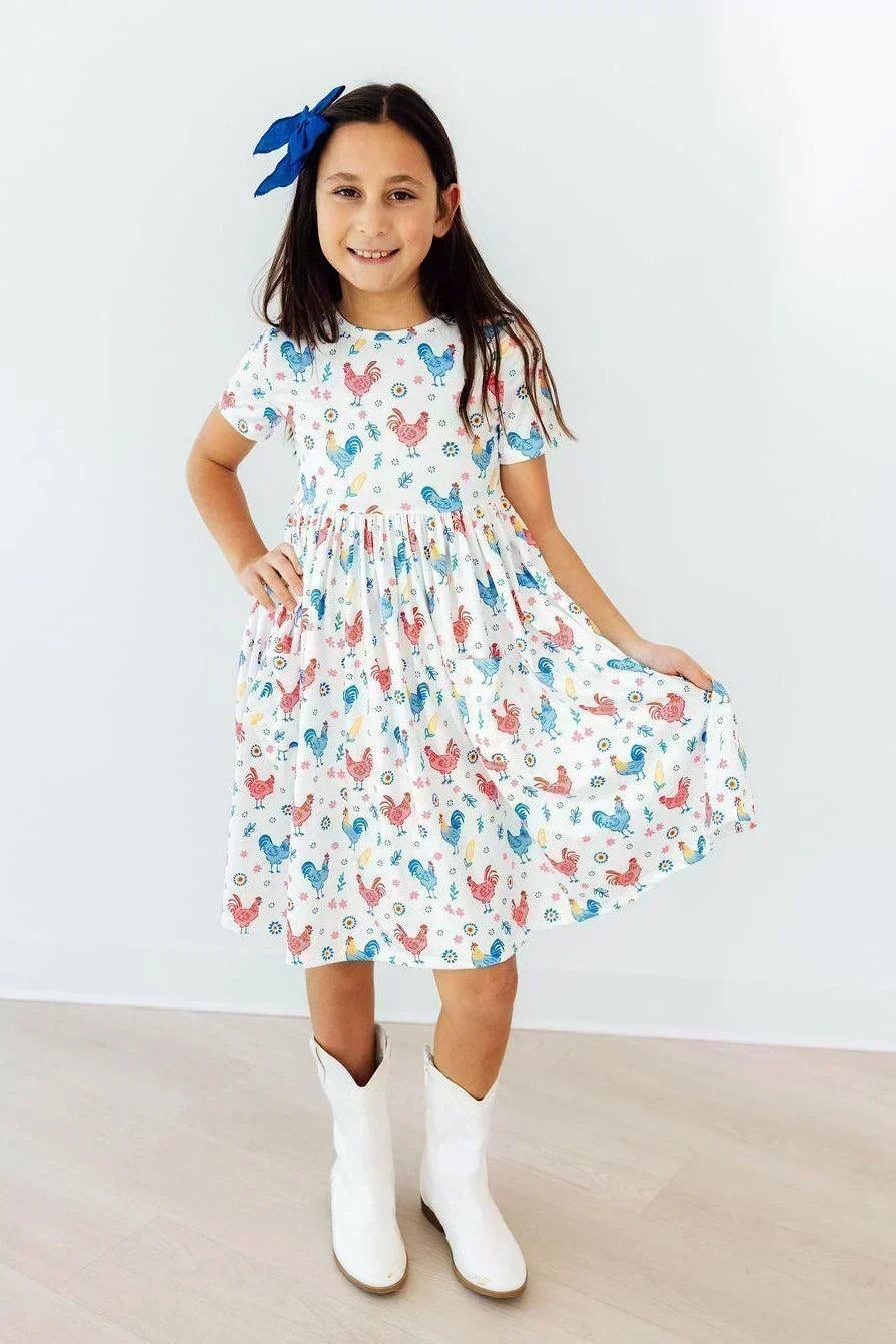 Young girl wearing a white short-sleeve dress with a colorful rooster print and white cowboy boots.