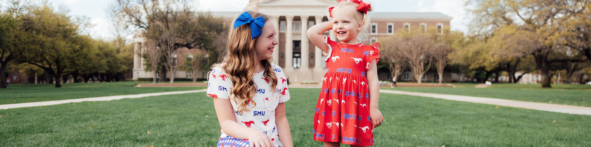 Two girls in SMU Mustangs apparel and hair bows playing on a grassy campus lawn in front of a historic brick building.