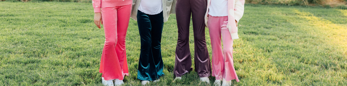 Four people wearing colorful velvet and corduroy bell bottom pants standing on a green grass lawn.
