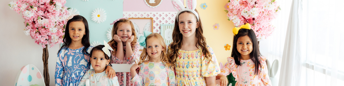 Three girls wearing pastel-colored Easter dresses with bunny, egg, and gingham prints in a festive spring setting.
