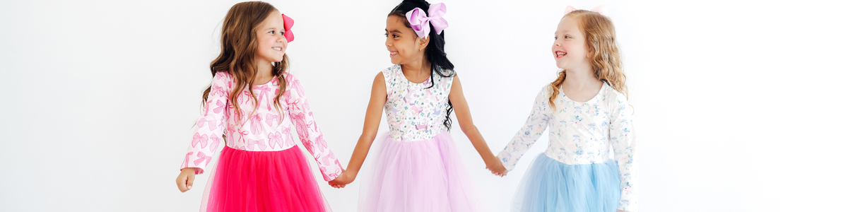 Three smiling girls wearing colorful tutu dresses with tulle skirts and patterned long-sleeve tops holding hands.
