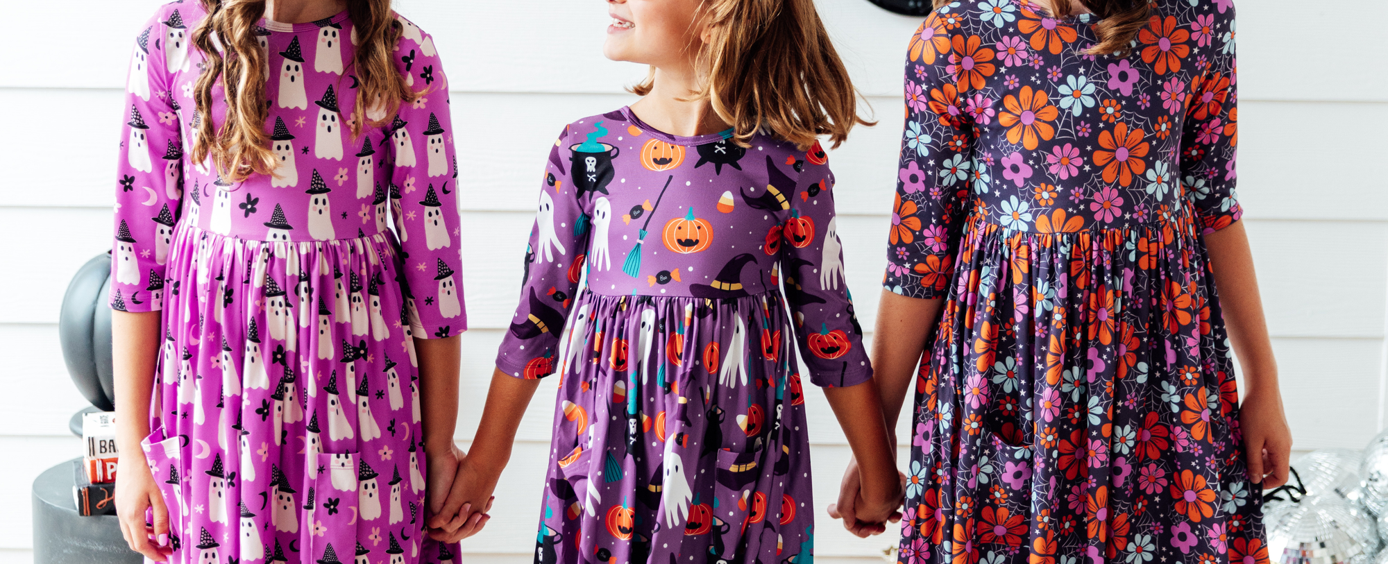 Three girls wearing purple long-sleeve Halloween dresses featuring ghost, pumpkin, and floral patterns.