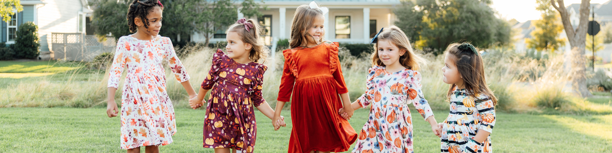 Five young girls holding hands in a grassy field wearing festive fall dresses with pumpkin and leaf prints.