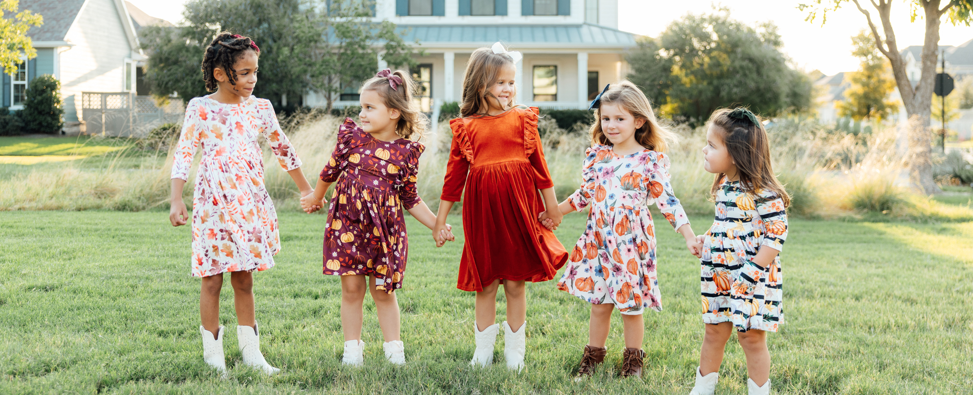 Five young girls holding hands in a grassy field wearing festive fall dresses with pumpkin and leaf prints.
