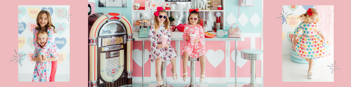 Two girls in heart sunglasses and patterned dresses holding pink milkshakes in a retro Valentine's Day themed diner.