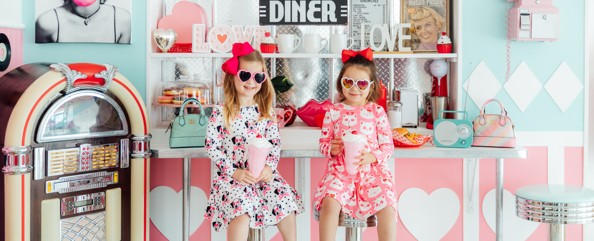 Two girls in heart sunglasses and patterned dresses holding pink milkshakes in a retro Valentine's Day themed diner.