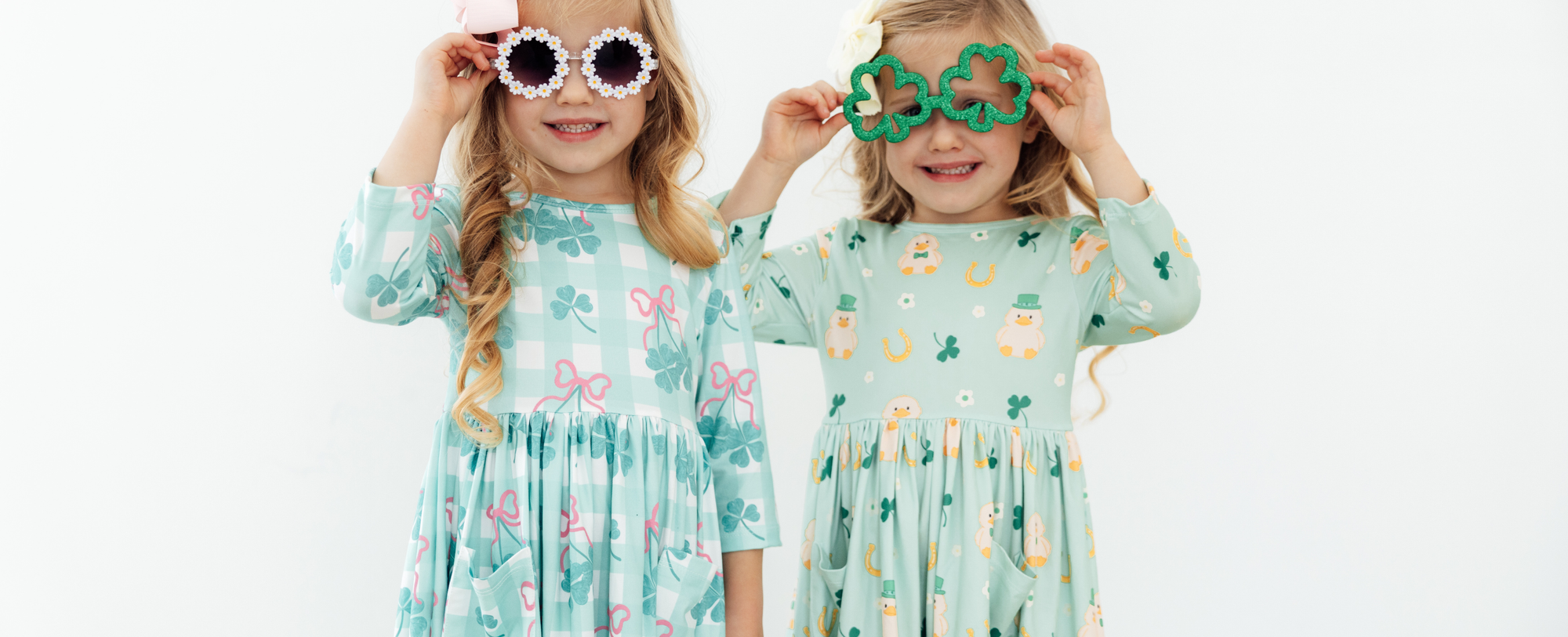 Two young girls in St. Patrick's Day dresses wearing festive daisy and shamrock-shaped sunglasses.