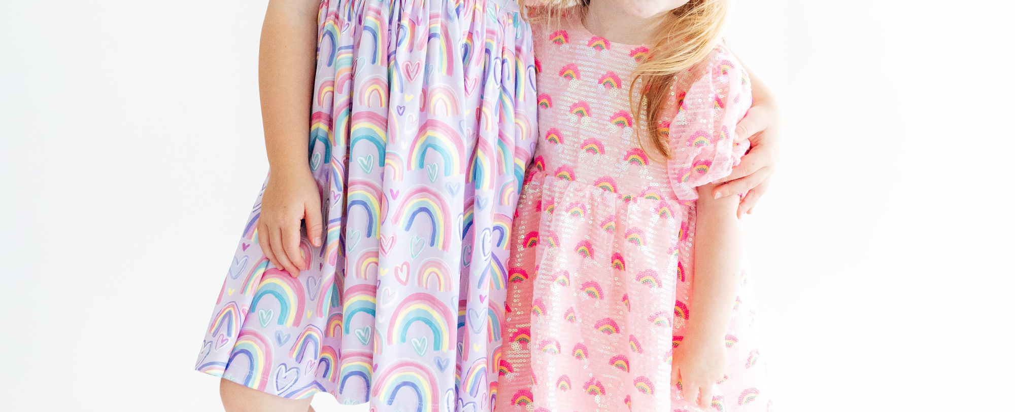 Two girls wearing colorful, playful dresses featuring rainbow and heart patterns against a white background.