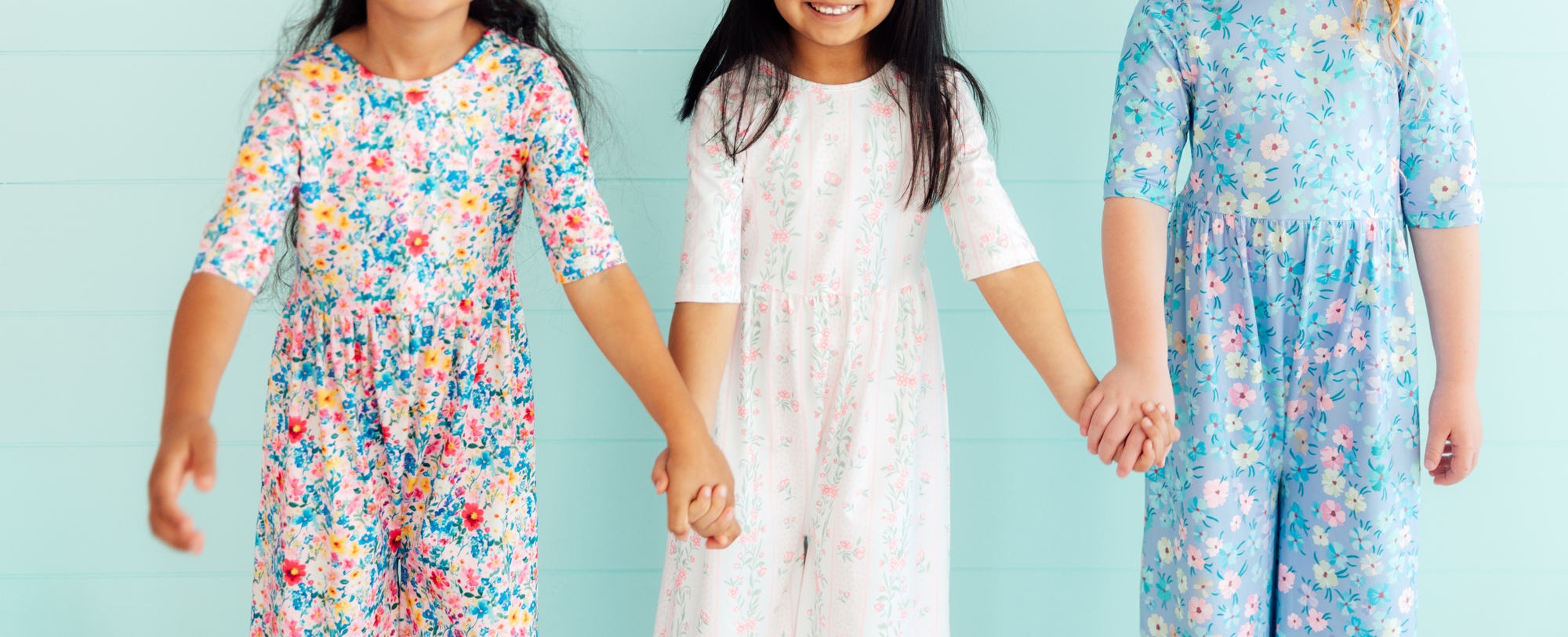 Three girls wearing colorful floral print wide leg jumpsuits standing against a light blue wooden wall.