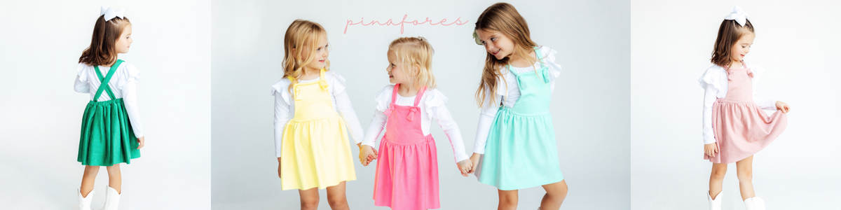 Four young girls smiling in colorful pastel pinafore dresses over white long-sleeve shirts in a classroom setting.