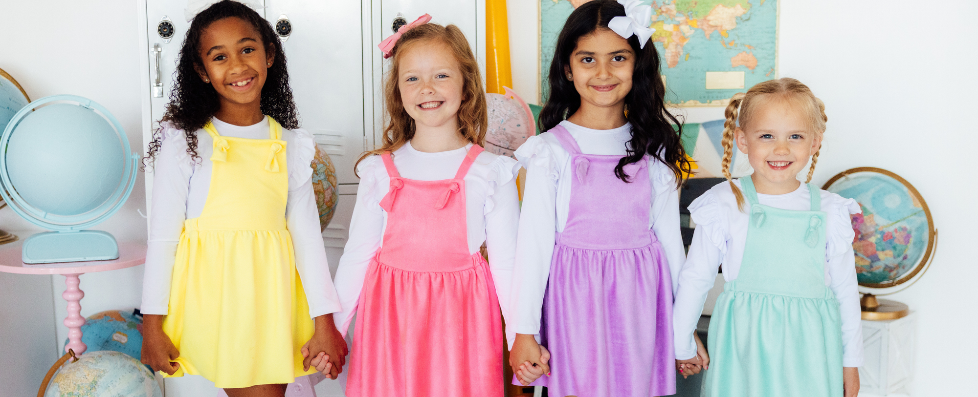 Four young girls smiling in colorful pastel pinafore dresses over white long-sleeve shirts in a classroom setting.