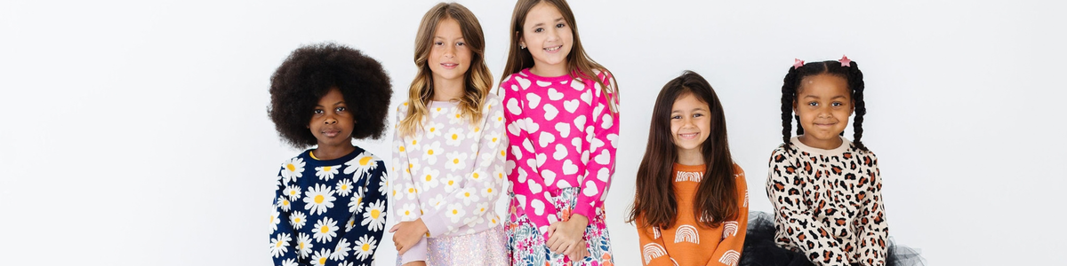Five smiling girls wearing colorful, patterned sweaters with daisy, heart, rainbow, and leopard prints.