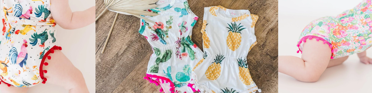 Baby rompers featuring colorful fruit and floral prints with playful pom-pom trim details on a wooden background.