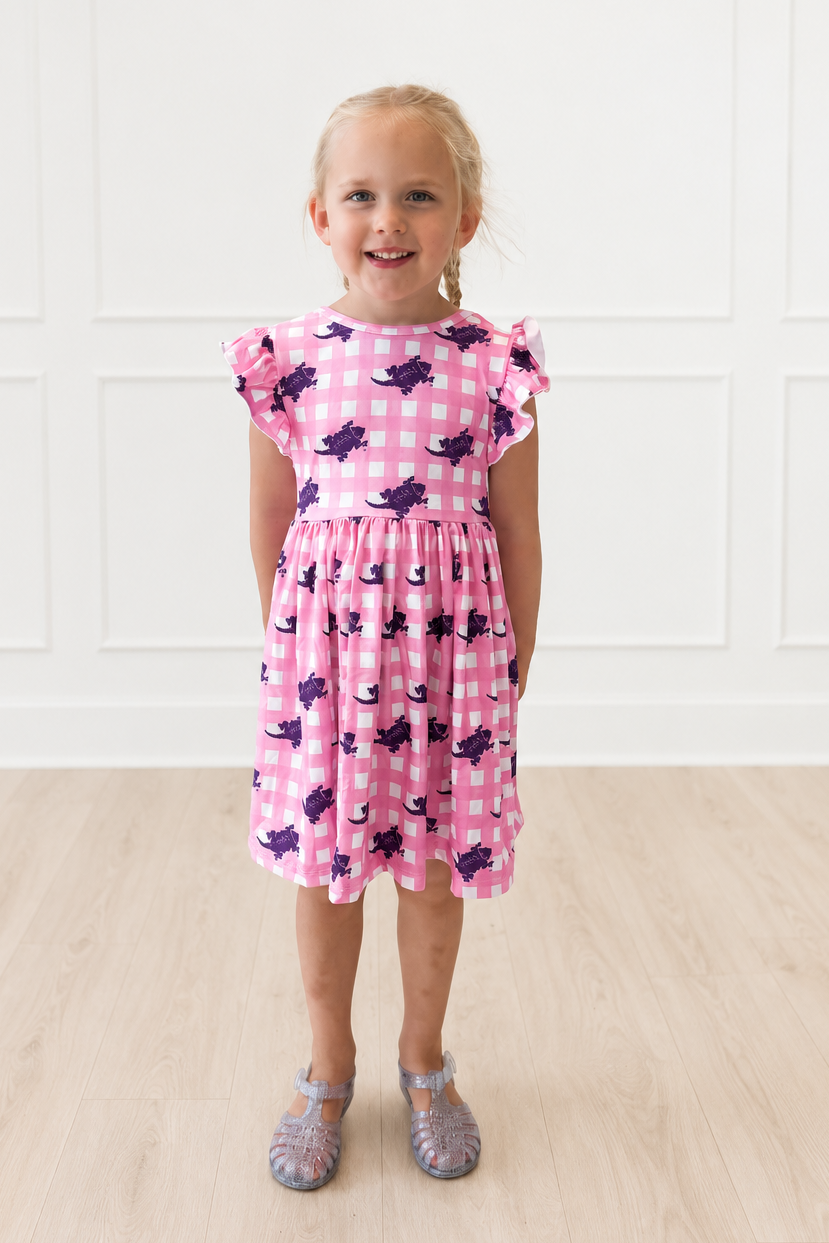 A young girl with blonde hair stands smiling in a pink and white gingham dress with purple Horned Frog print, paired with sparkly silver shoes, set against a white paneled wall and light wood floor.