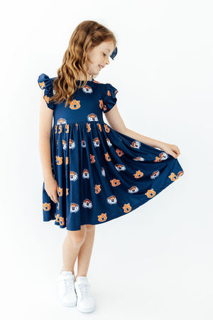 A young girl with long wavy light brown hair is wearing a navy blue dress with a colorful cartoon bear pattern, holding out her skirt to show the fabric's flow. She is standing against a seamless white studio background.