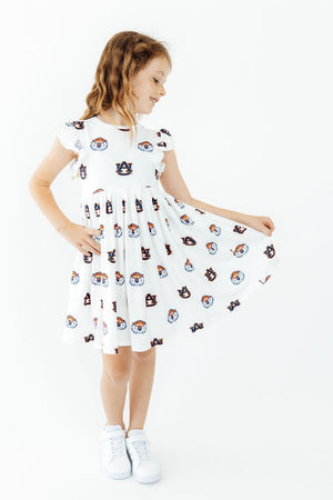 A young girl with light brown hair stands against a white studio background, wearing a white short-sleeve dress with a colorful cartoon character print and white sneakers, holding out the skirt to show its volume.