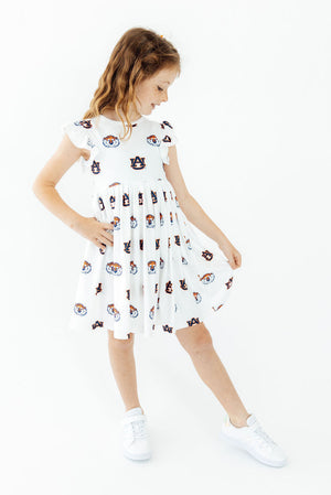 A young girl wearing a white short-sleeve dress with a black and multicolor cartoon face pattern, paired with white sneakers, standing against a plain white background.