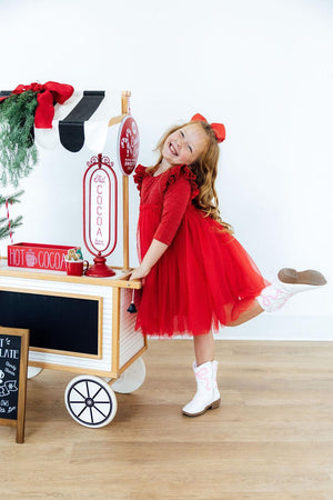 A young girl in a red holiday dress and white cowboy boots poses next to a toy hot cocoa cart on a light wood floor against a white wall.